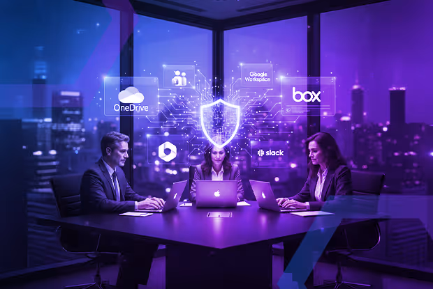 Three business professionals working on laptops at a table with a digital shield and technology icons floating above representing cybersecurity and data protection.