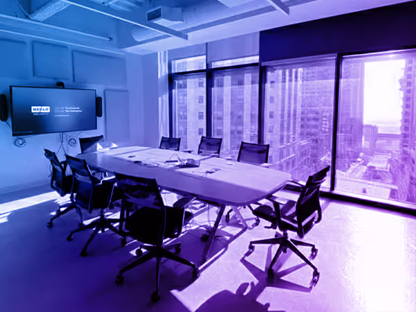 Modern conference room with a large table surrounded by black office chairs, a wall-mounted screen, and floor-to-ceiling windows overlooking a cityscape.