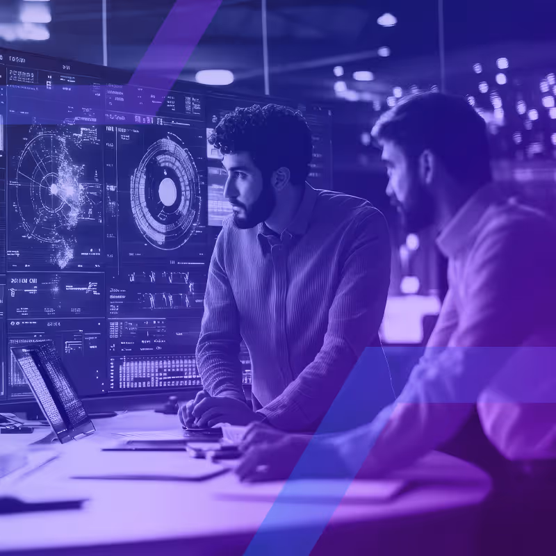 Two men analyzing complex data and digital charts on large computer screens in a dimly lit modern office.