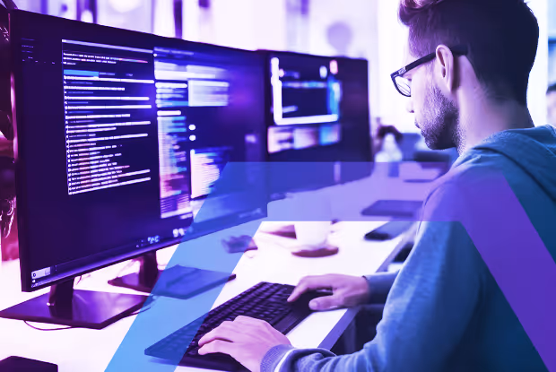 Man wearing glasses working at a desk with two large monitors displaying code and digital interfaces.