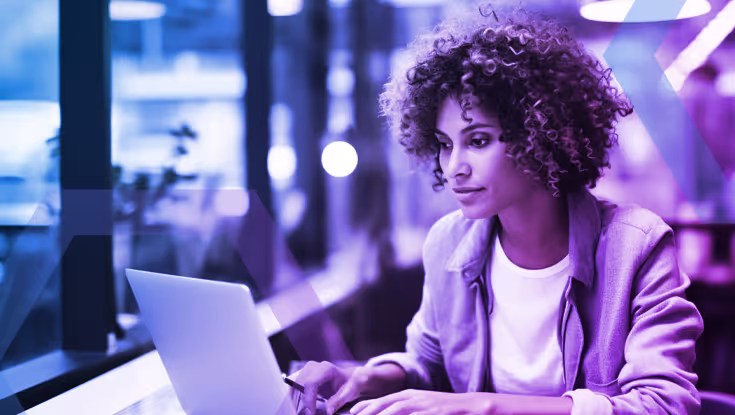 Woman with curly hair using a laptop at a desk near a window in a modern office setting.