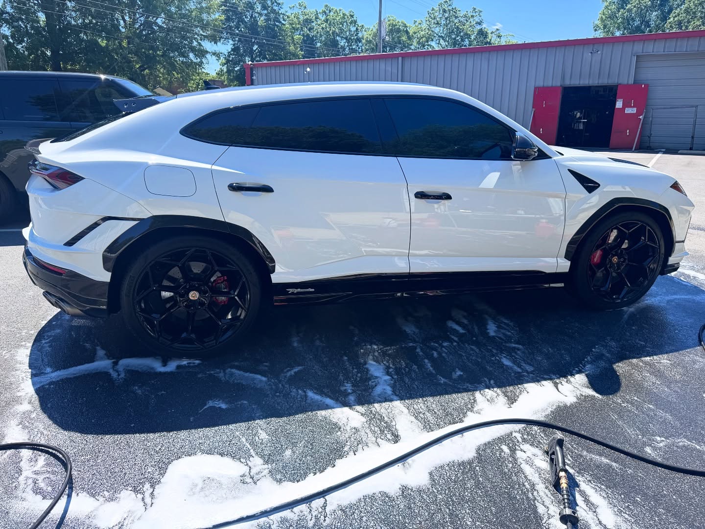White Lamborghini SUV with black wheels parked on a soapy driveway near a building with red doors.