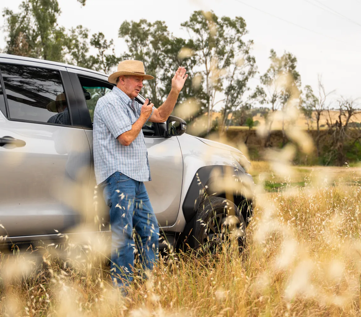Older man in a straw hat using a walkie-talkie while standing next to a silver pickup truck in a grassy field with trees in the background.