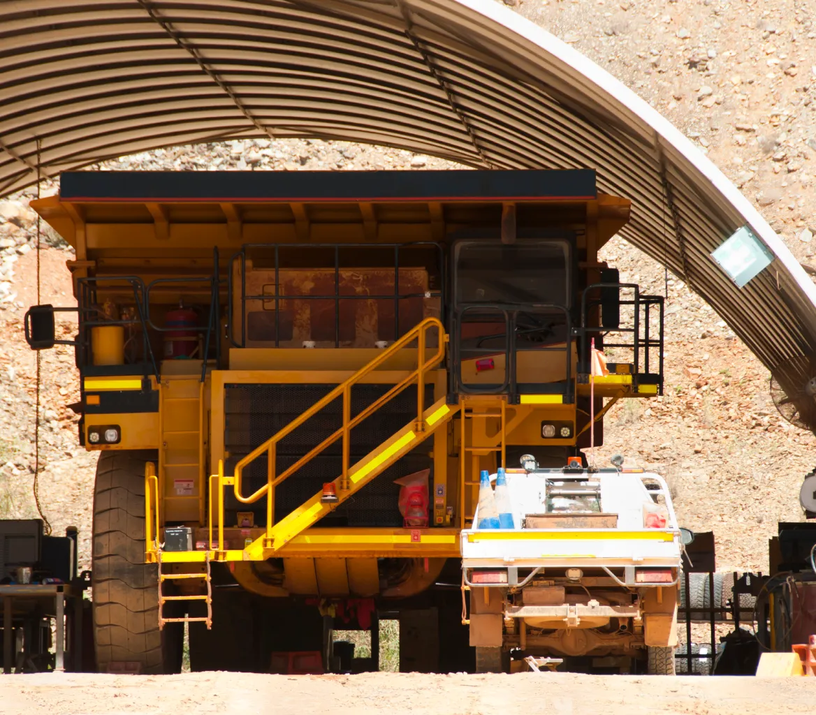 Large yellow mining dump truck and smaller white utility vehicle parked under a corrugated metal shelter in a rocky mining area.