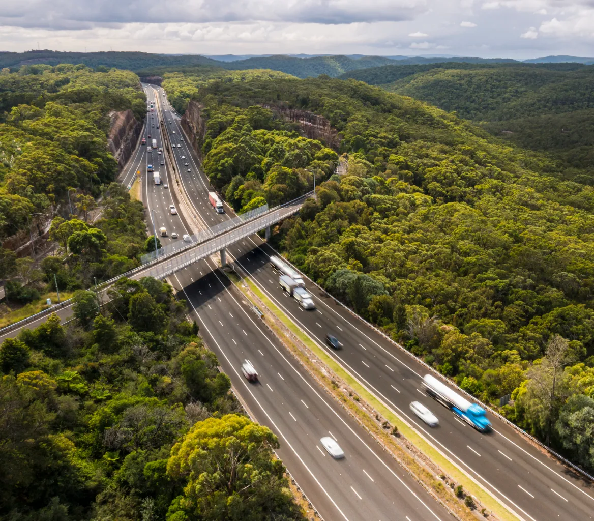 Aerial view of a busy highway cutting through dense green forest with a pedestrian bridge crossing over it.