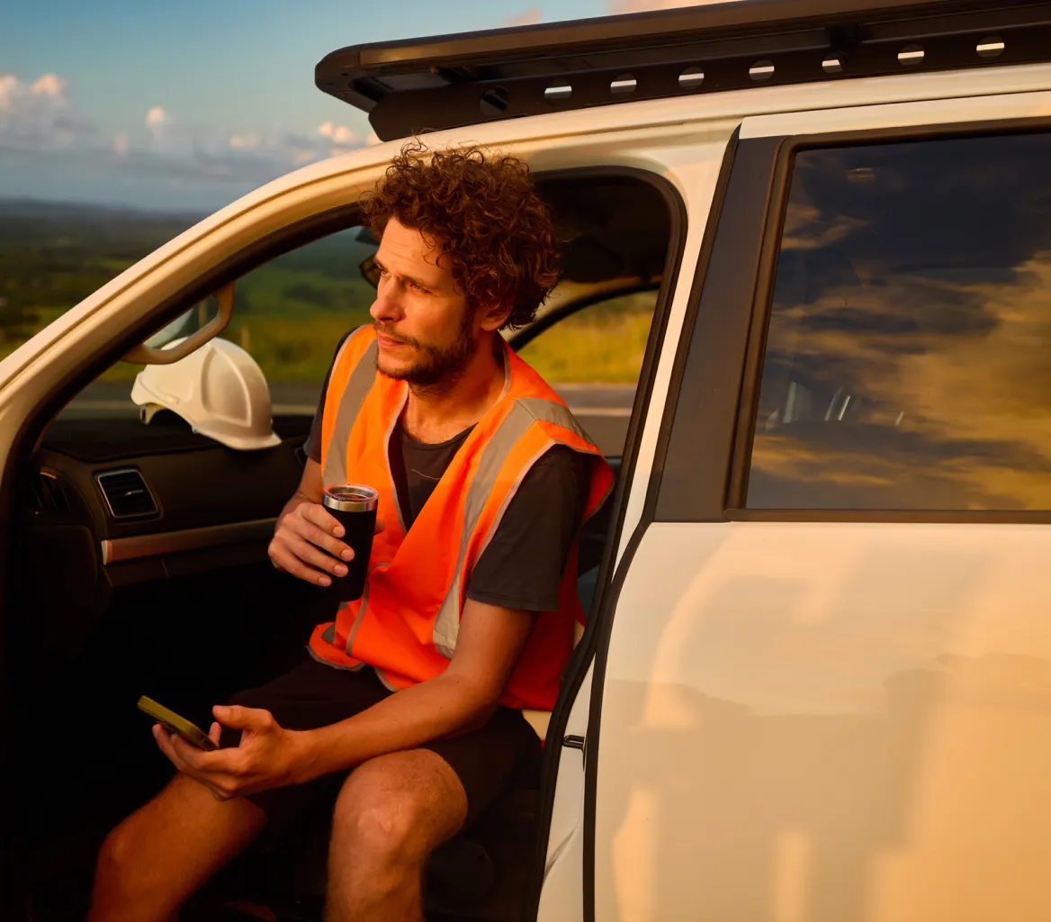 Man in an orange safety vest sitting in a white vehicle with the door open, holding a phone and a drink.