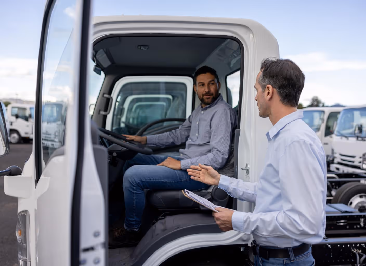 Man sitting inside a white truck cab listening to another man standing outside, holding a clipboard, with multiple trucks in the background.