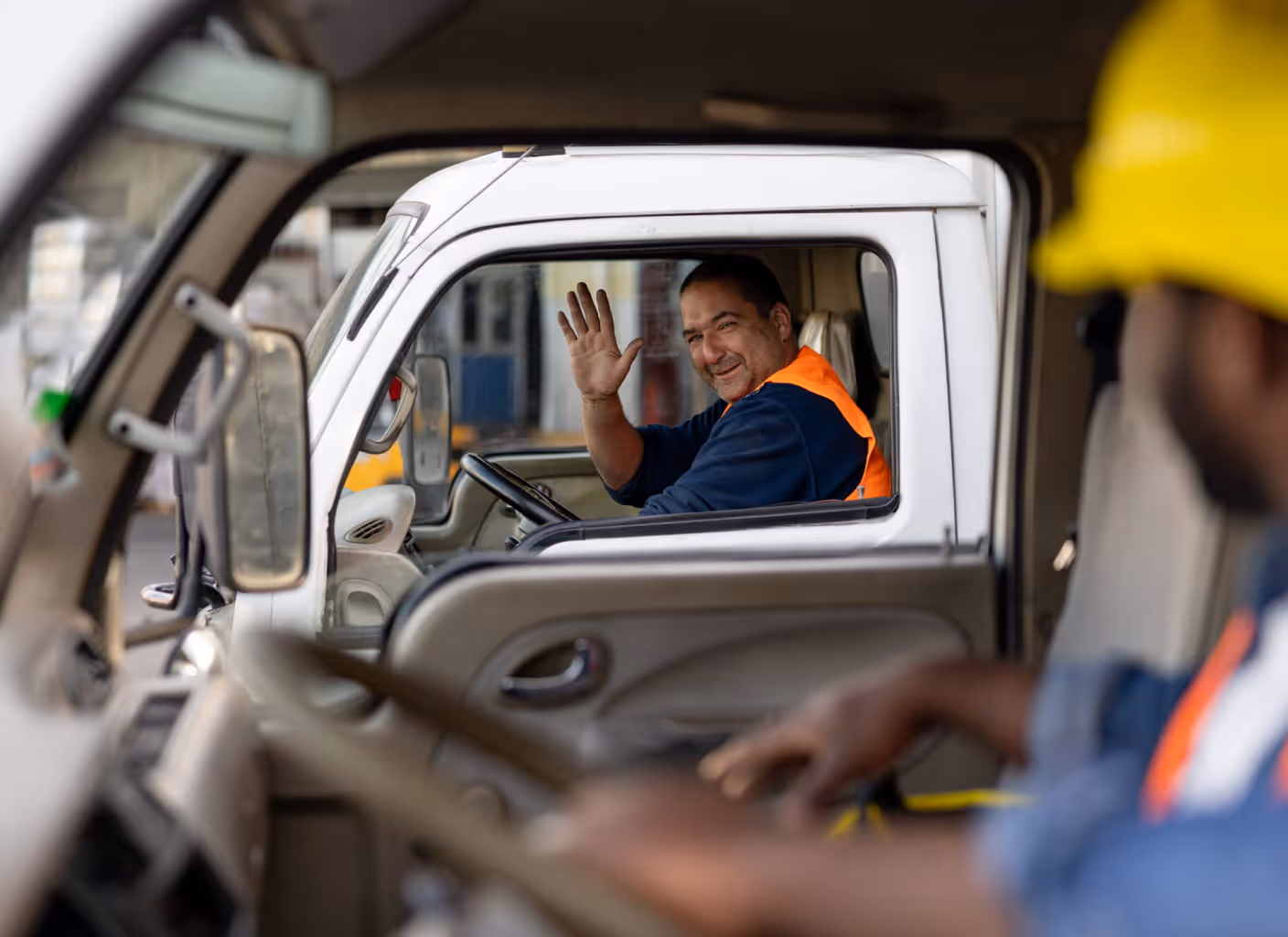Smiling man in a white truck waving through the open window wearing a dark shirt and orange safety vest, another person wearing a yellow helmet seen blurred in foreground.