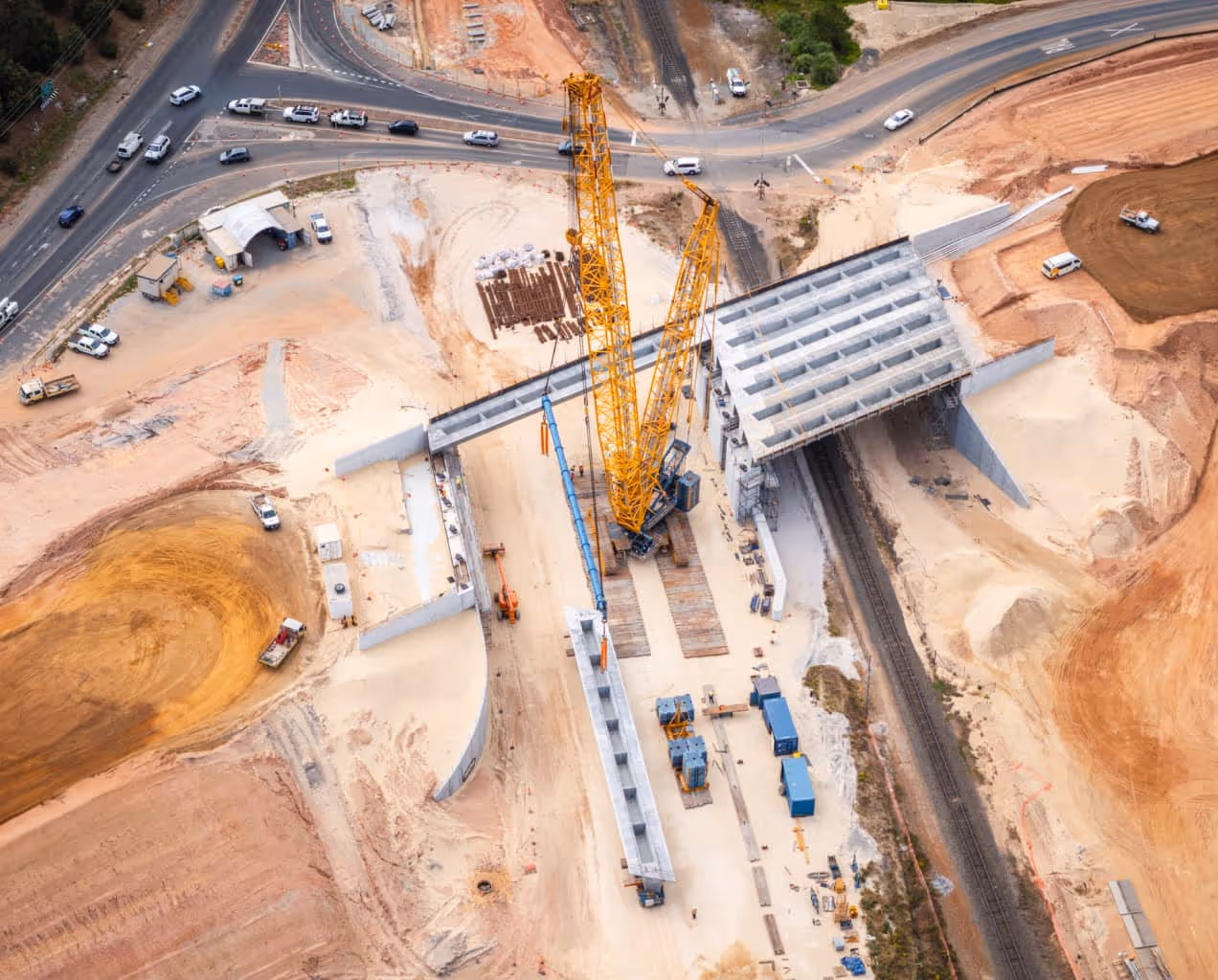 Aerial view of a construction site with large yellow cranes assembling concrete bridge segments over railway tracks.