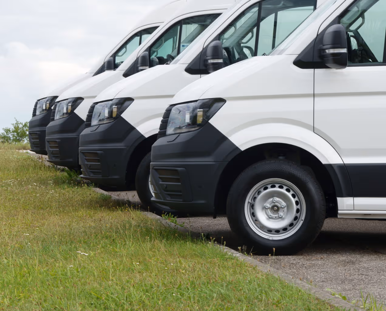 Row of white commercial vans parked next to a grassy area under a cloudy sky.