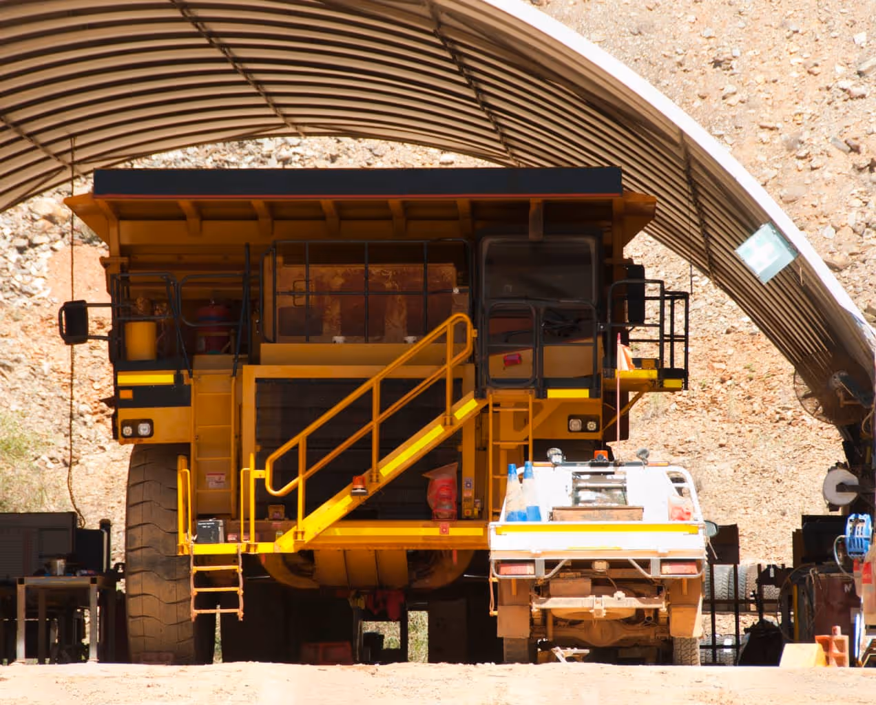 Large yellow mining dump truck and smaller utility vehicle parked under a curved metal shelter at a mining site.
