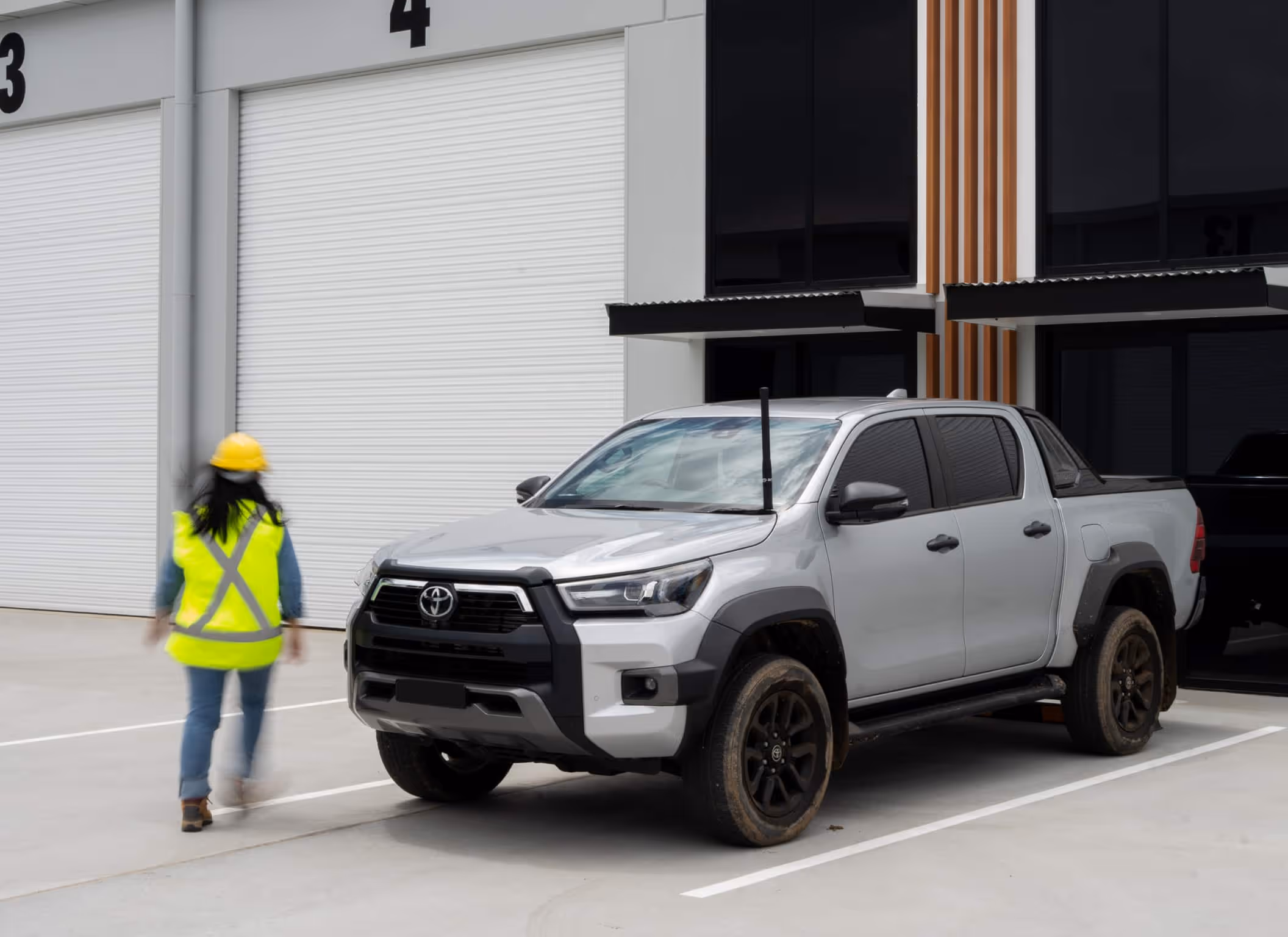 Silver Toyota pickup truck parked outside a warehouse with a person in a yellow safety vest and helmet walking nearby.