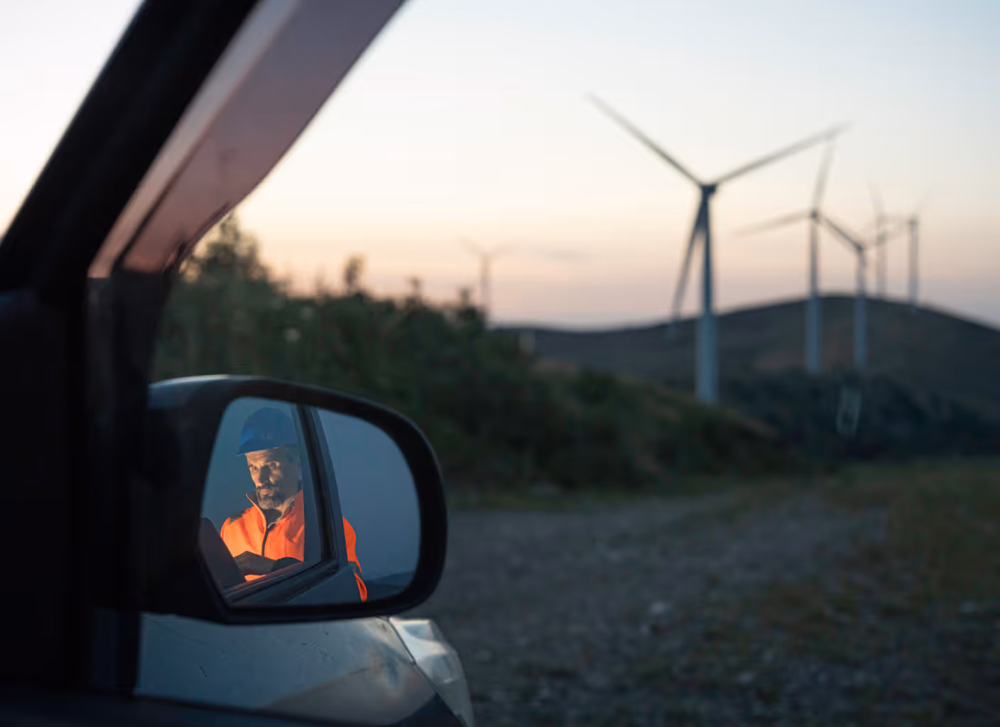 Reflection of a man in an orange safety jacket and blue helmet looking at a device in a car's side mirror with wind turbines in the background at dusk.