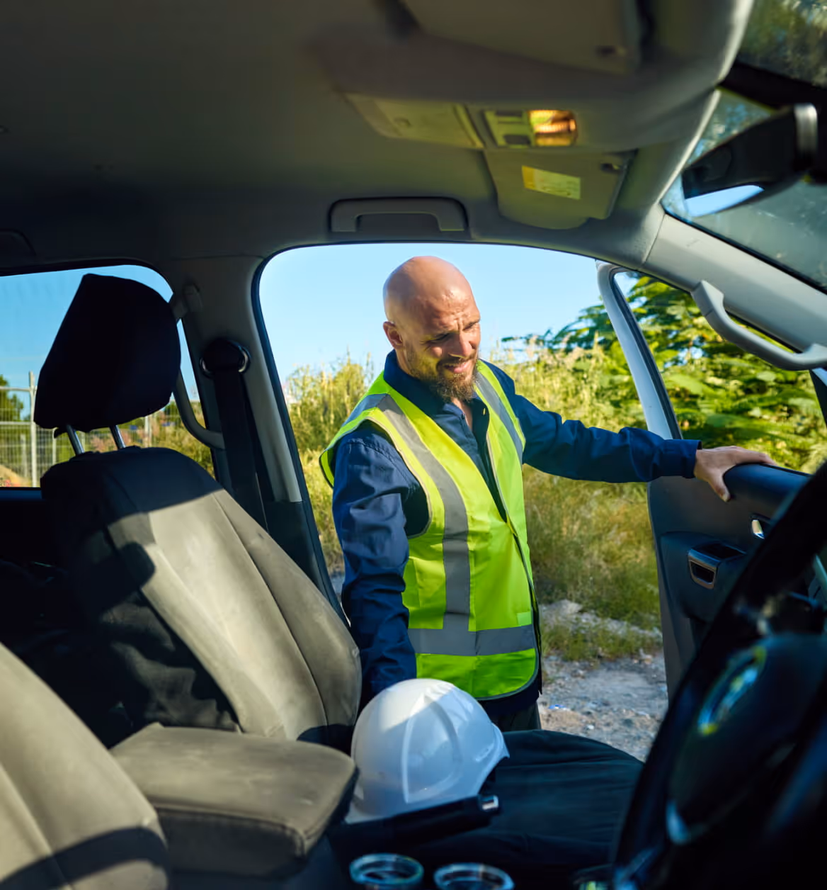 Man wearing a yellow safety vest standing next to an open car door, holding the door while a white construction helmet rests on the front passenger seat.