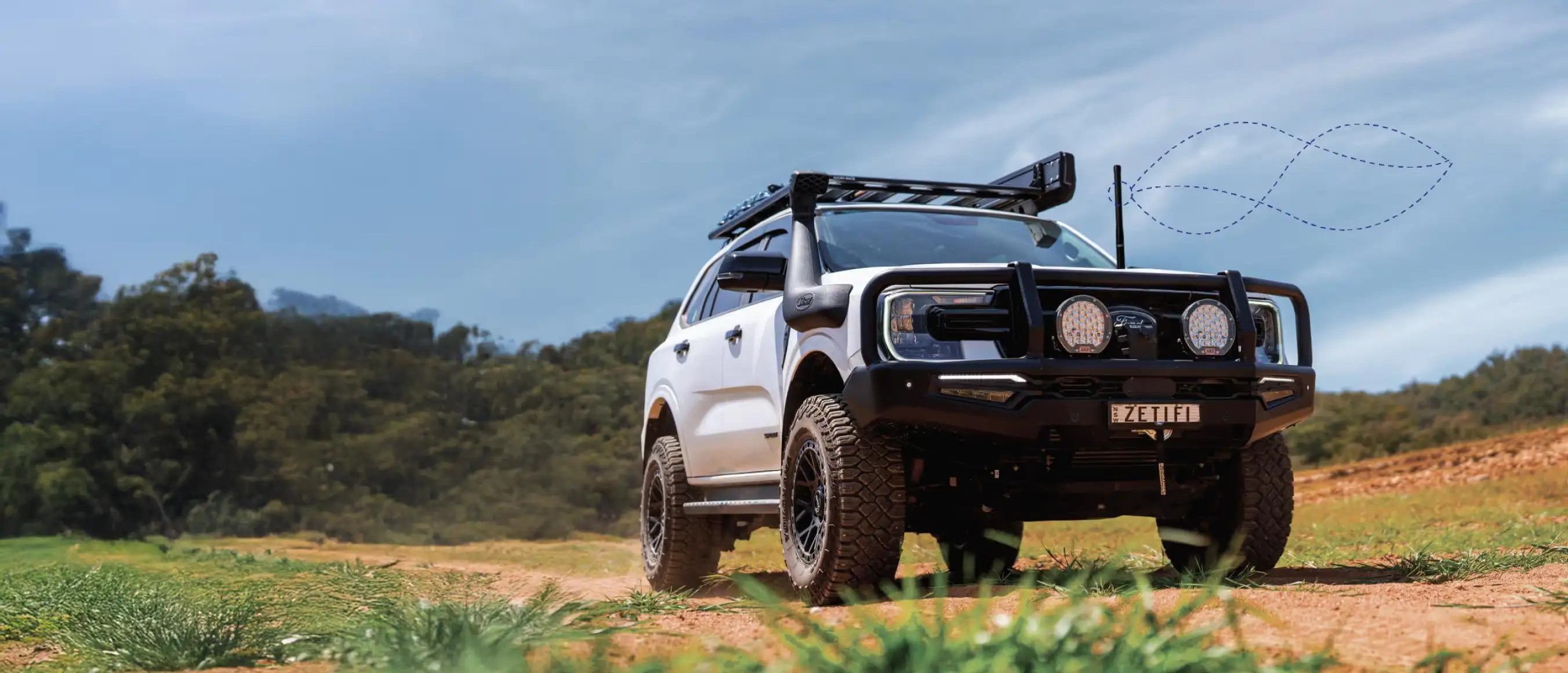White off-road SUV equipped with roof rack, snorkel, and front bull bar on a dirt path with green vegetation and blue sky background.