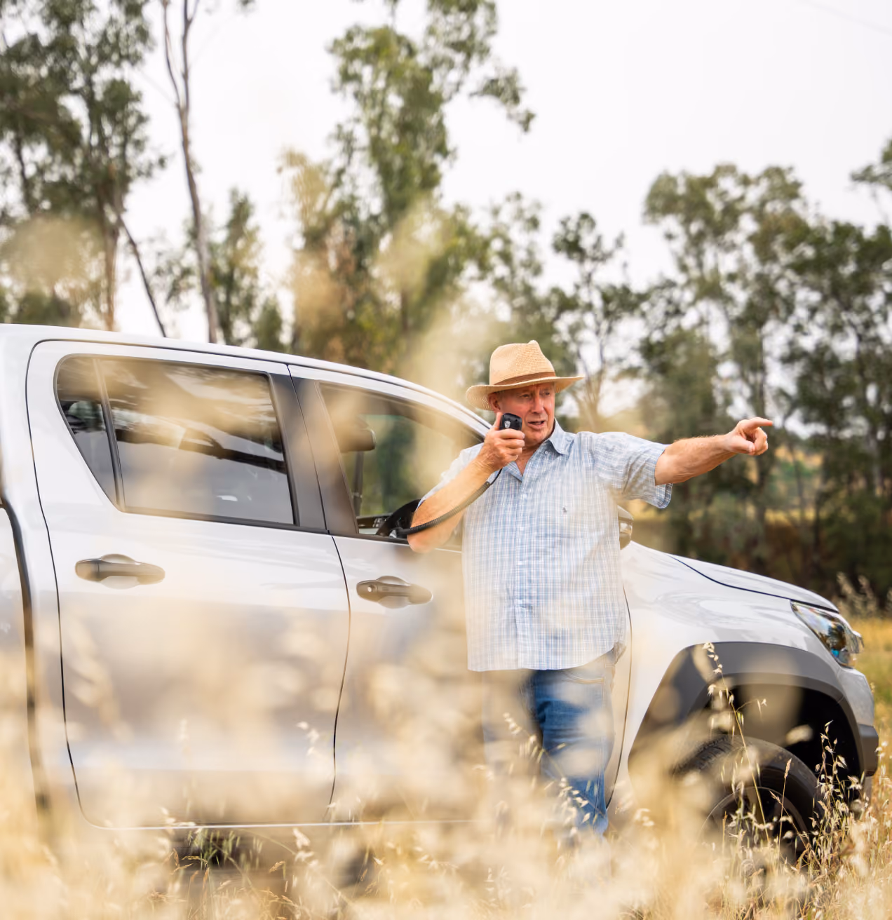 Man in straw hat standing by white pickup in a field, speaking into a handheld radio and pointing.