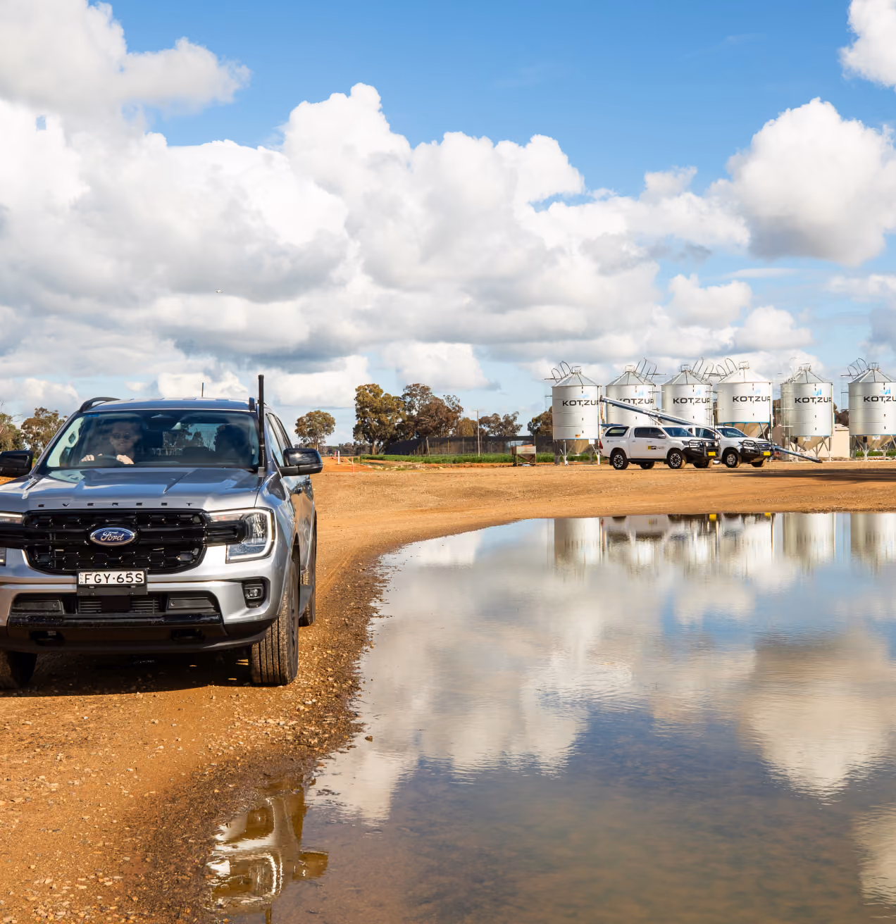 Silver Ford pickup on dirt road beside large water puddle reflecting sky and grain silos in background.