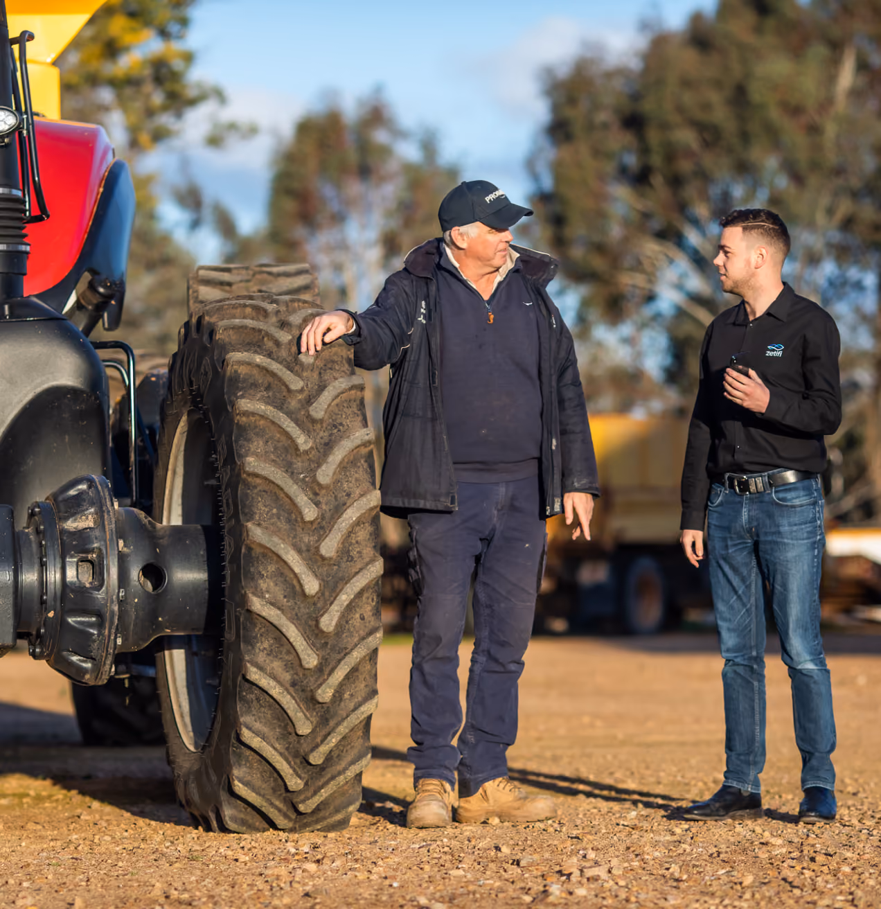 Two men standing near a large tractor tire outdoors, engaged in conversation under clear sky.