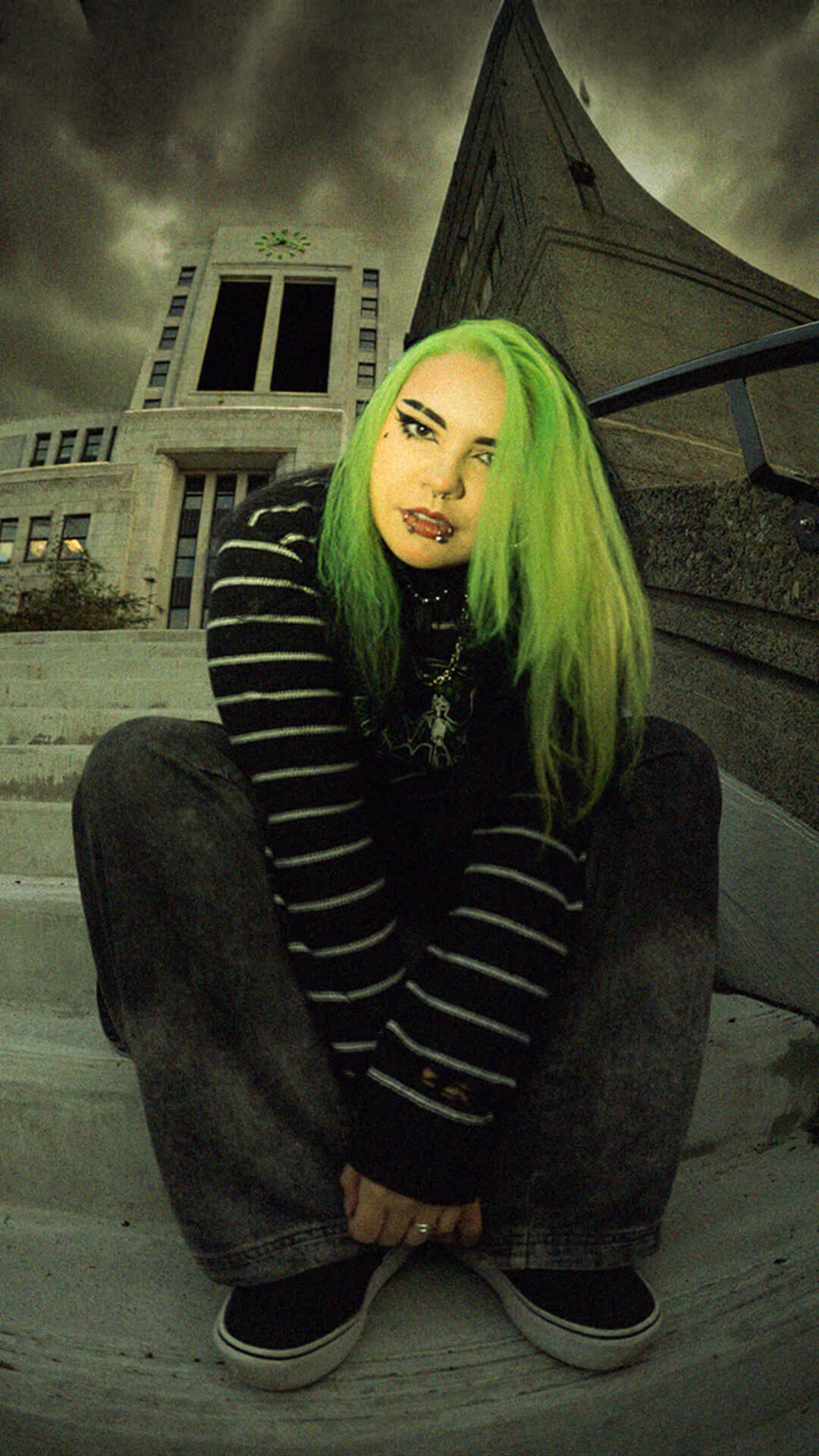 Young woman with red hair, wearing neon green sunglasses, black shirt with green logo, and dark makeup, standing in front of an escalator.