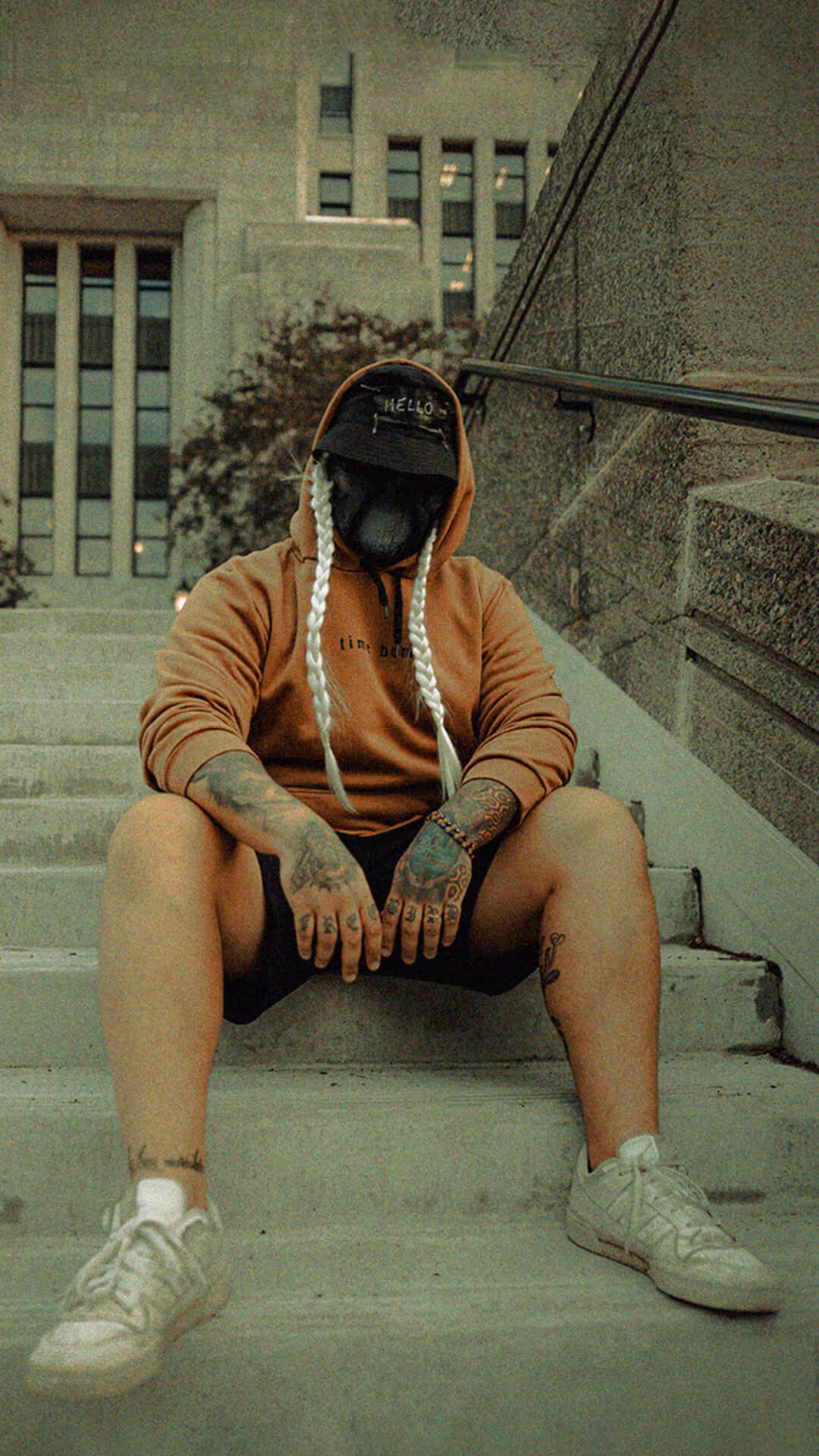 Person with long white braided hair wearing a black bucket hat with 'HELLO' on it, a black face mask, black shirt, and tattooed arms leaning against a green tiled wall.