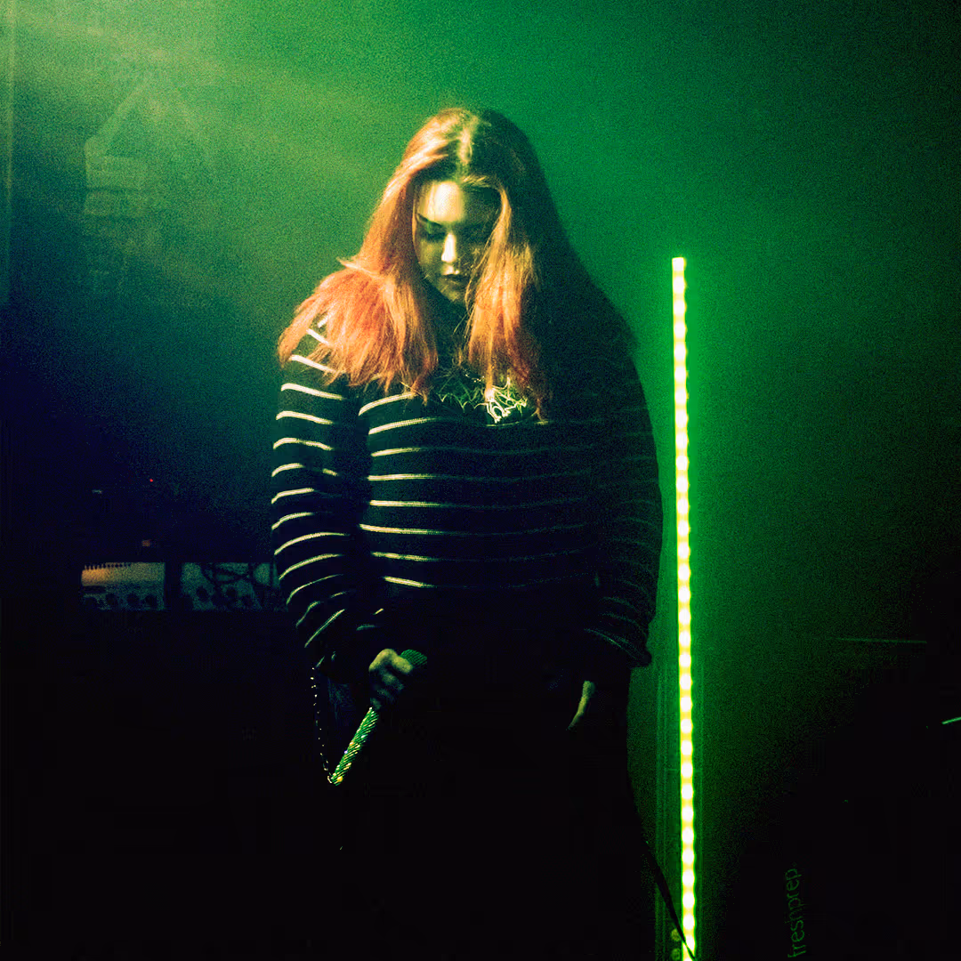 Red-haired vocalist passionately singing into a microphone on stage with a drum set and graffiti art of a large bird in the background.