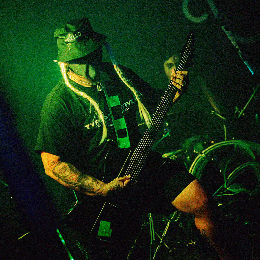 Musician playing electric guitar on stage with drummer in background and colorful graffiti on wall.