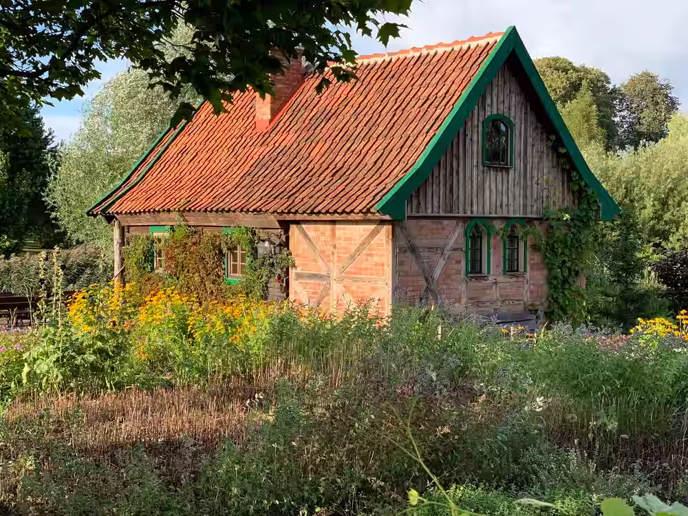Drewniano-ceglany domek z czerwonym, ceramicznym dachem i zielonymi obramowaniami okien oraz dachu, otoczony bujną roślinnością i kwitnącymi żółtymi kwiatami, położony wśród drzew w letnim, słonecznym świetle.