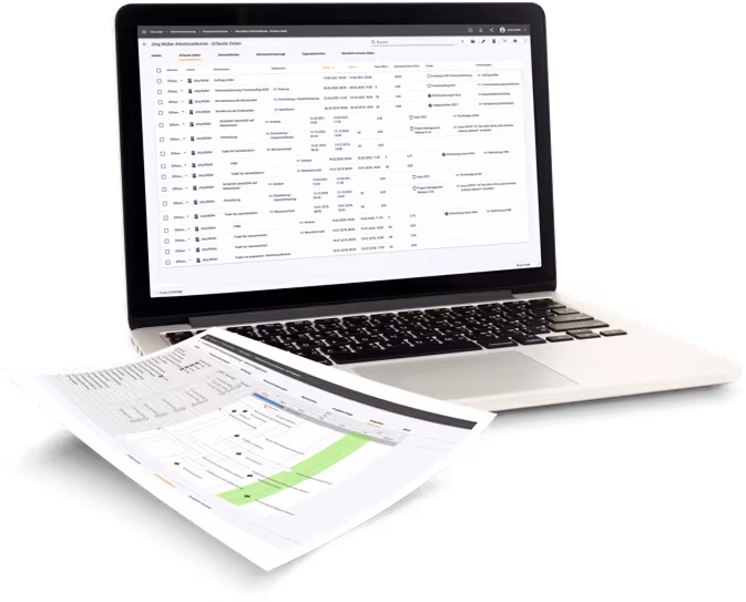 Open laptop displaying a detailed table of work data in German, with a printed spreadsheet showing a project timeline or workflow chart placed in front of it.