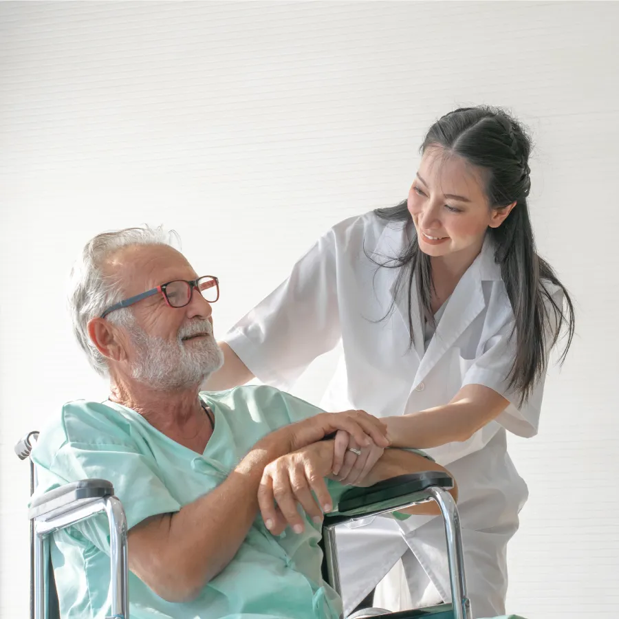 Smiling female healthcare worker holding hands with elderly male patient seated in wheelchair.