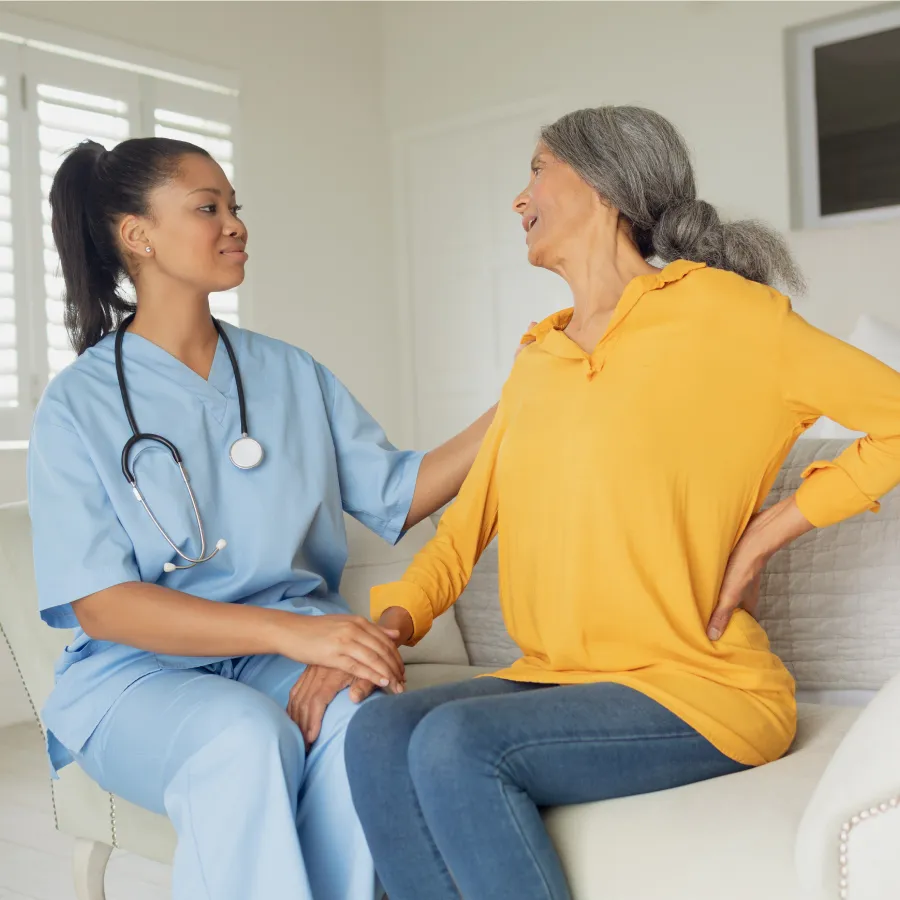 Nurse in blue scrubs comforting an elderly woman in a yellow sweater sitting on a couch.