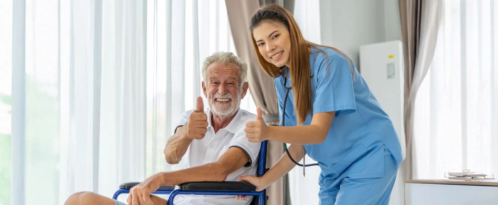 Smiling elderly man in a wheelchair giving a thumbs-up with a female nurse in blue scrubs beside him also giving a thumbs-up.