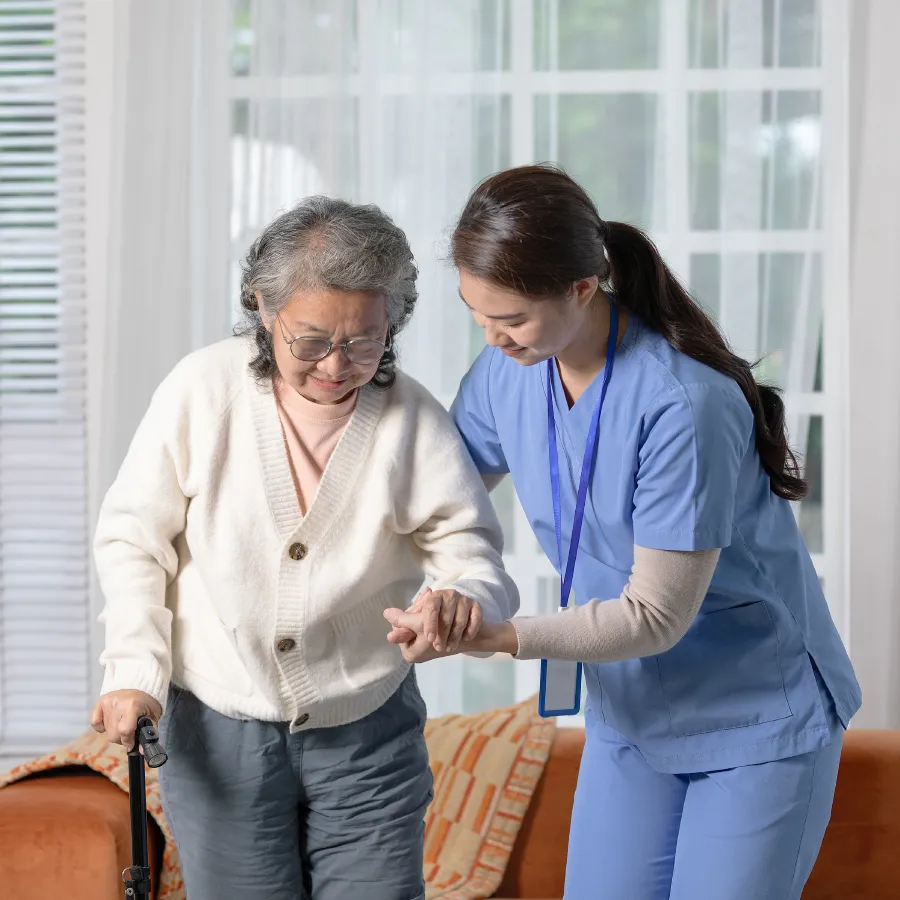 Healthcare worker in blue scrubs assisting elderly woman with glasses and walking cane.