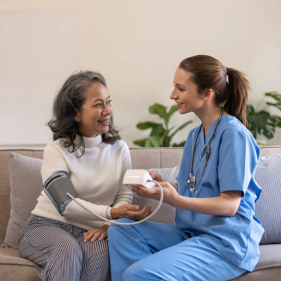 Nurse in blue scrubs smiling and checking blood pressure of elderly woman sitting on a couch.