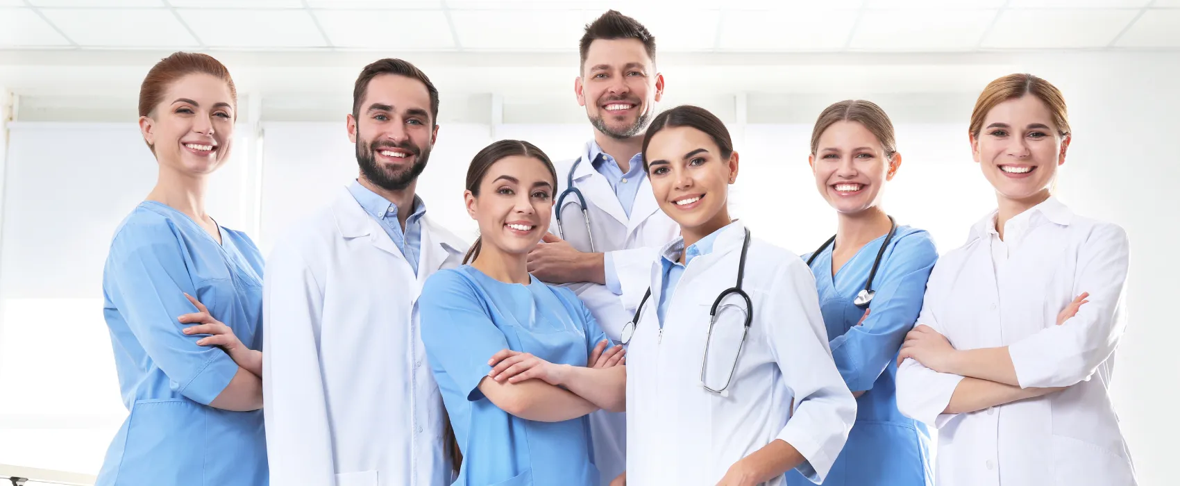 Group of seven smiling diverse medical professionals in scrubs and lab coats standing with arms crossed in a bright clinic room.