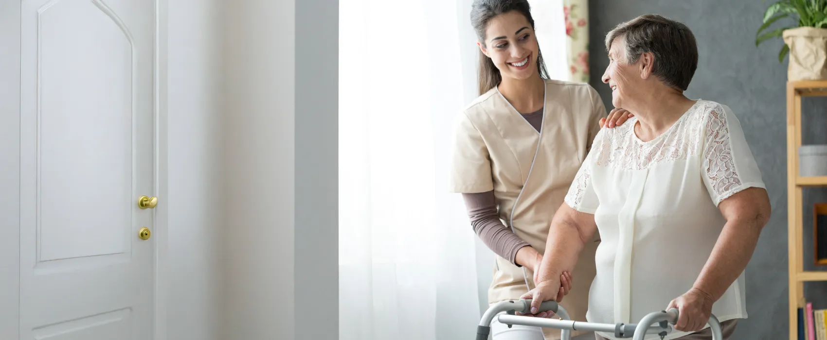 Caregiver smiling and assisting an elderly woman using a walker inside a bright room.