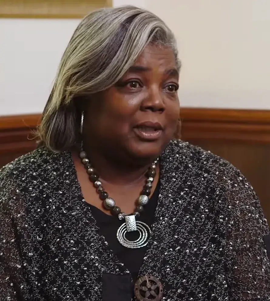 Middle-aged woman with gray and black hair wearing a black patterned sweater and a statement necklace speaking indoors.