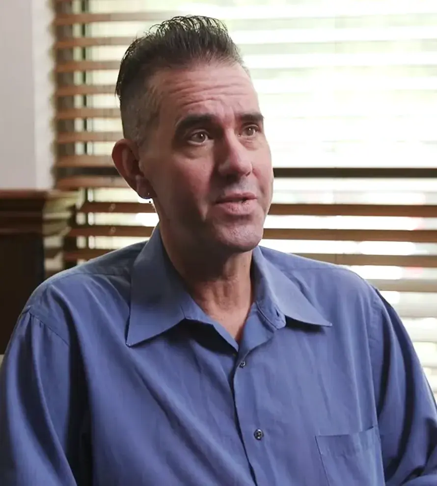 Man with slicked-back dark hair and an earring, wearing a blue button-up shirt, seated indoors with wooden blinds in the background.