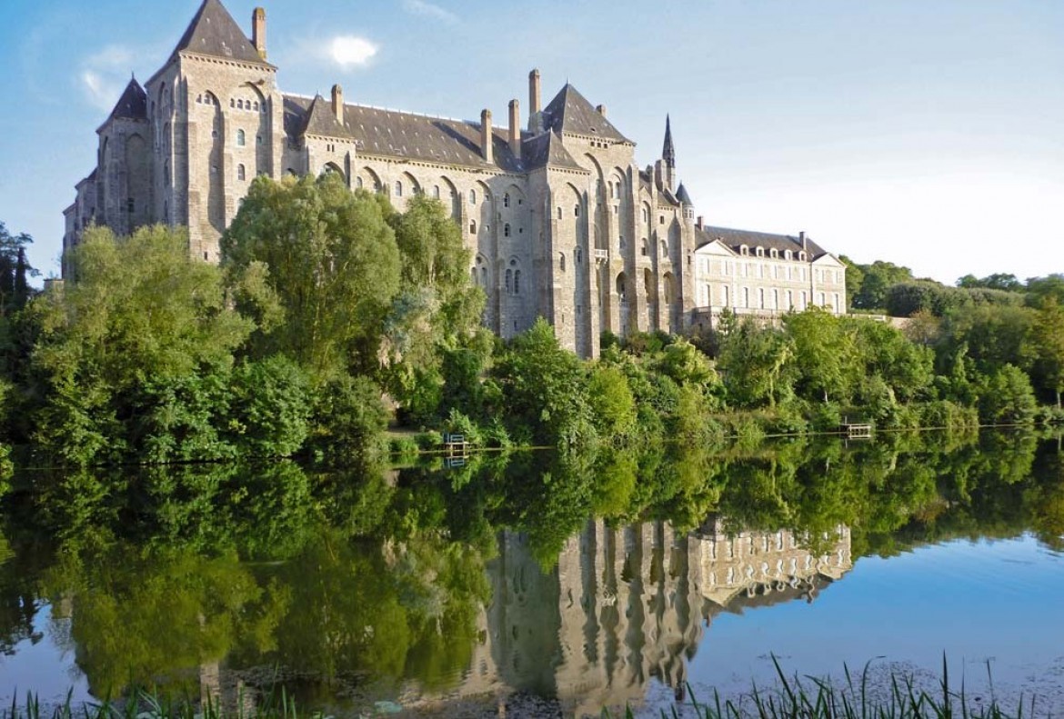 Large historic stone abbey with multiple towers and pitched roofs, surrounded by dense green trees reflected in a calm river.