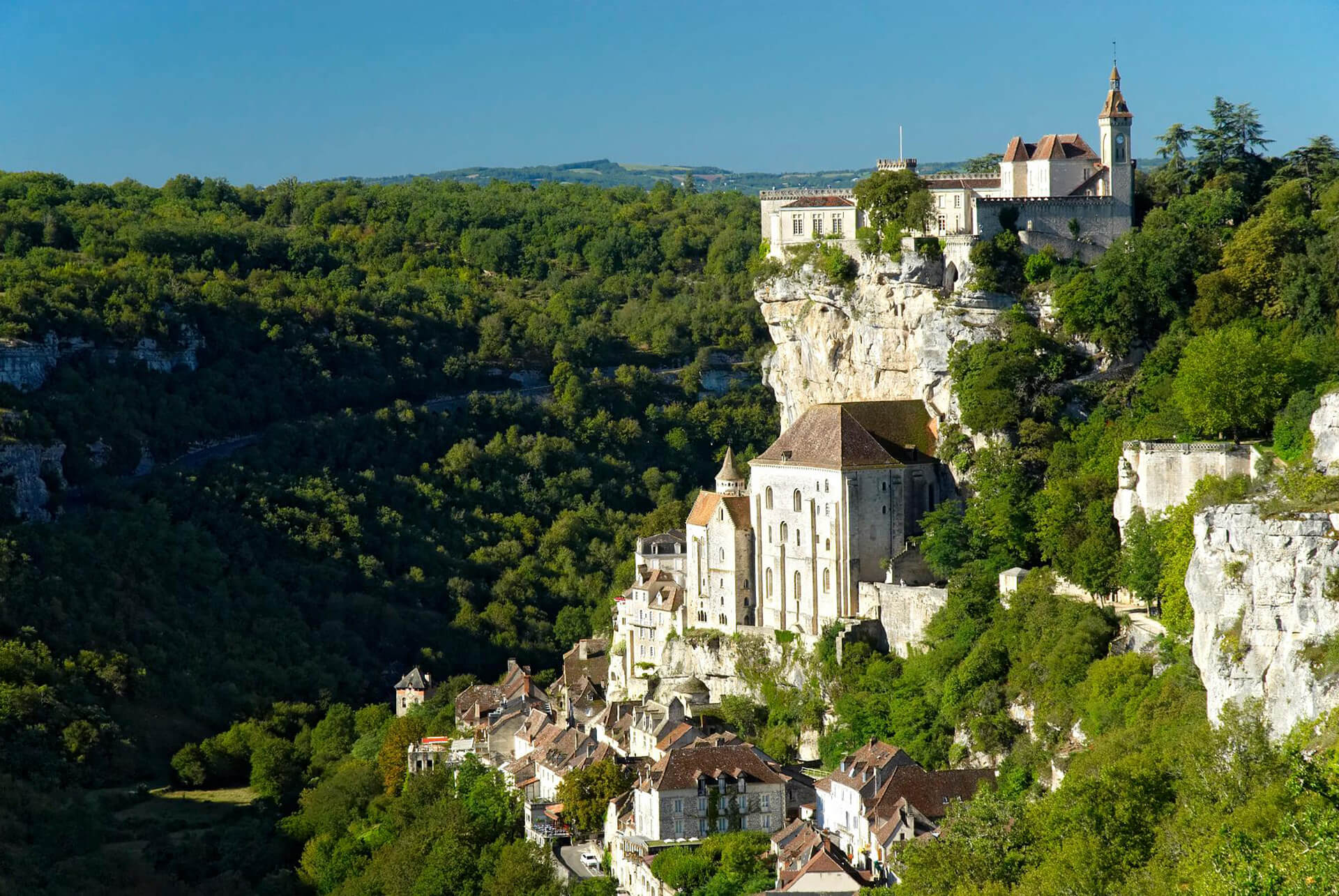 Medieval village with stone buildings and a large church perched on a cliff surrounded by dense green forest under a clear blue sky.