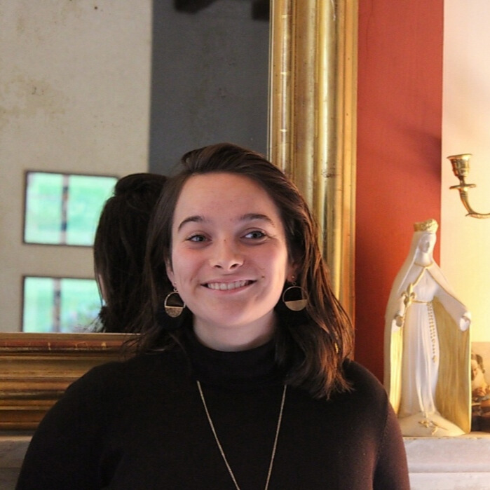 Smiling woman with shoulder-length dark hair wearing a black turtleneck and gold earrings standing indoors in front of a large gold-framed mirror and a statue on a mantle.
