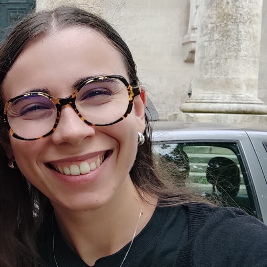 Smiling woman with tortoiseshell glasses and silver earrings outdoors near a car and stone column.