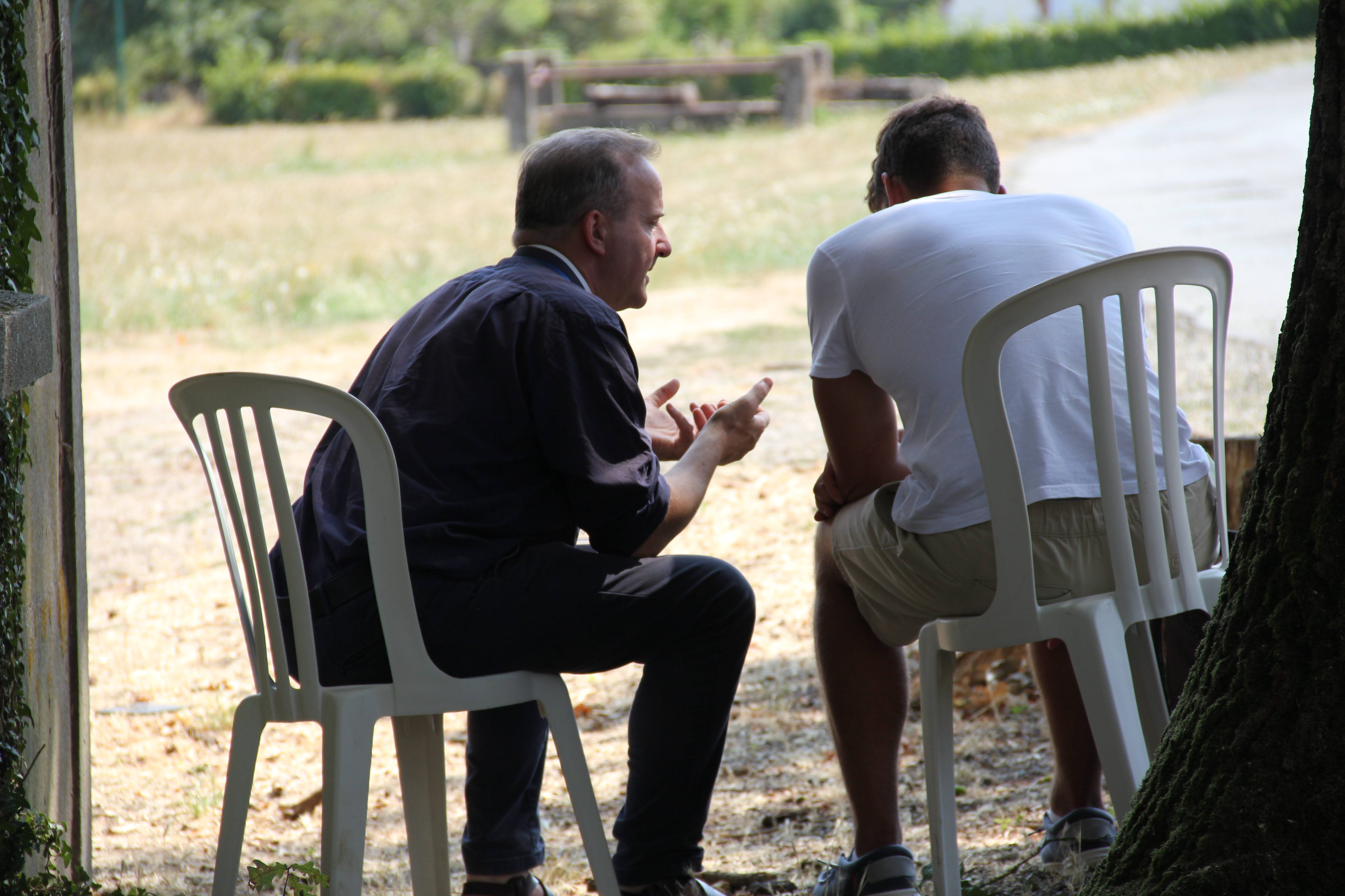 Two men sitting on white plastic chairs outdoors, engaged in a conversation; one man wearing a dark shirt is gesturing with his hands, the other is in a white t-shirt and shorts, their backs mostly to the camera with a blurred natural background.