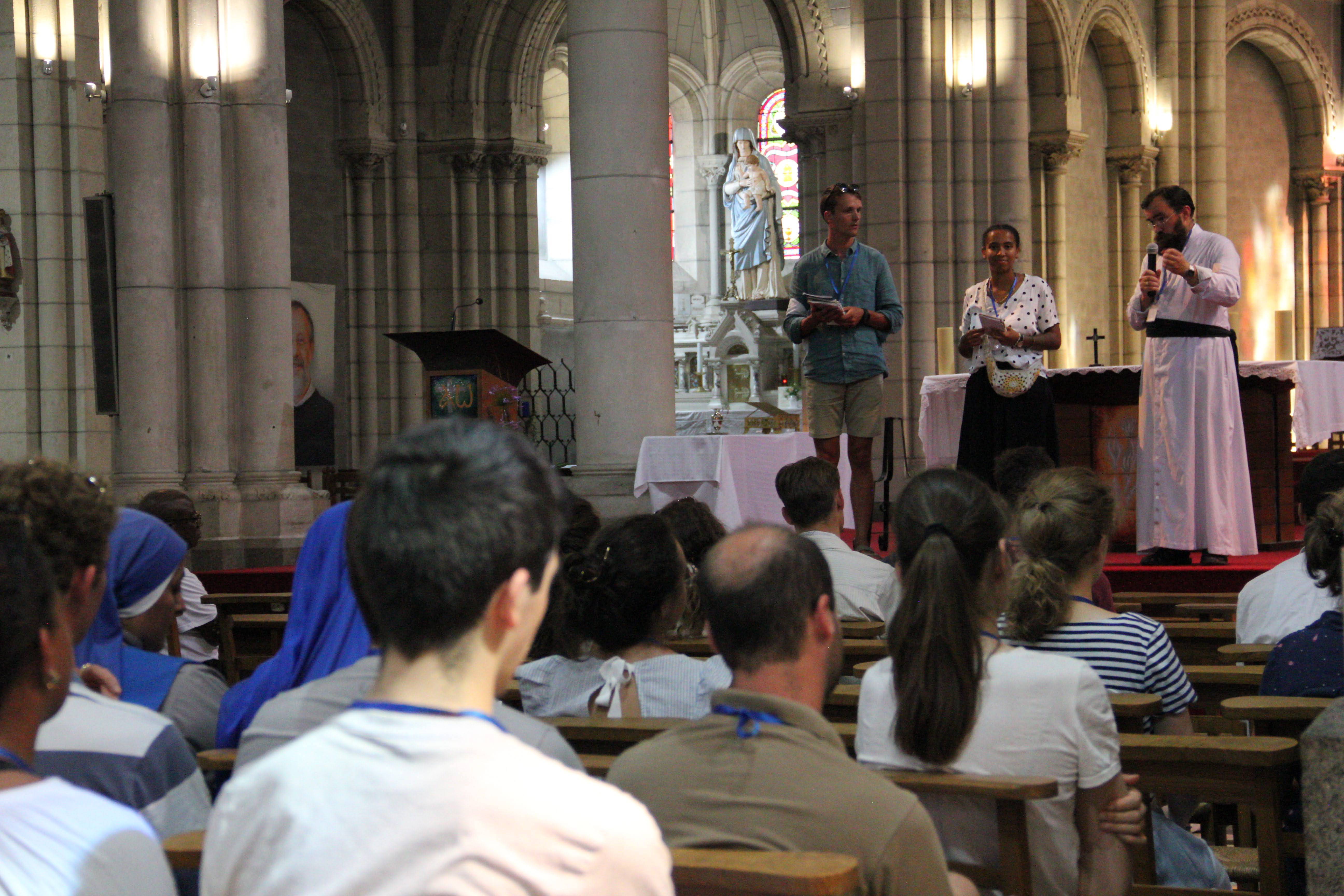People seated in wooden benches inside a large church with arched ceilings, facing a priest in white robes speaking into a microphone and two individuals holding papers standing near the altar.