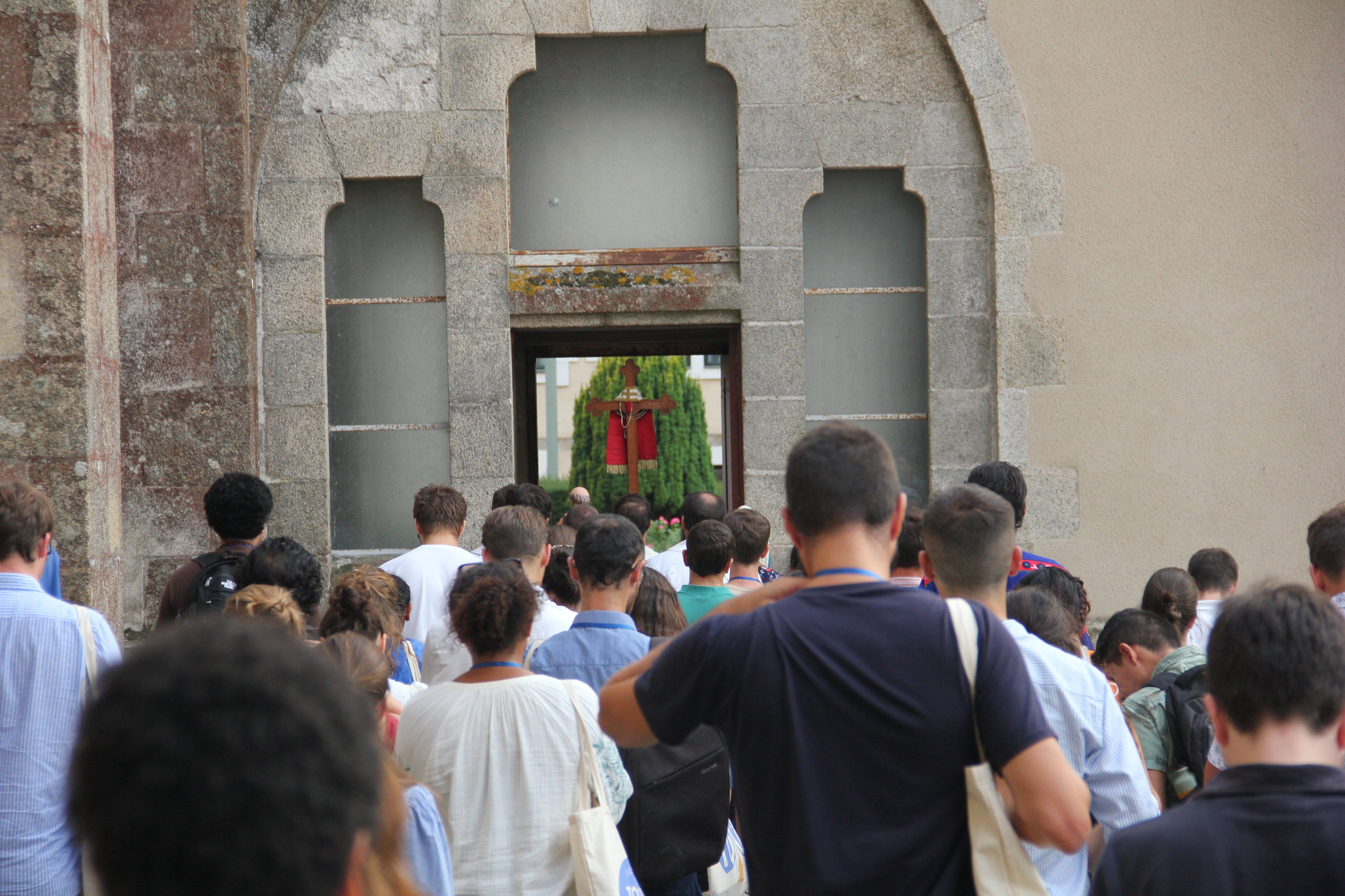 Group of people seen from behind walking toward a stone archway with a wooden cross draped with red cloth visible through the opening.
