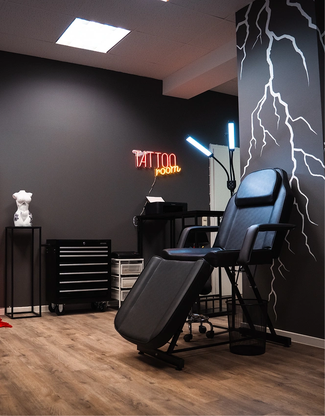 Tattoo room with black chair, illuminated neon sign, lightning wall art, and drawers on wood floor.