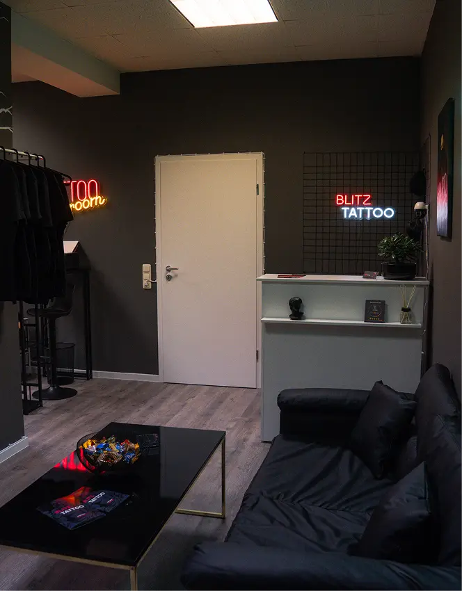 Tattoo studio waiting area with black couch, coffee table, clothes rack, neon signs, and reception desk.
