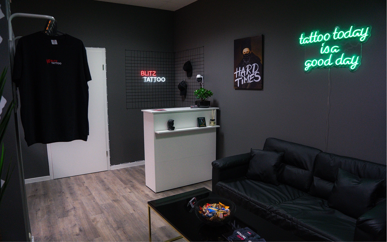 Tattoo shop interior with black couch, white counter, neon signs, and a hanging black T-shirt.