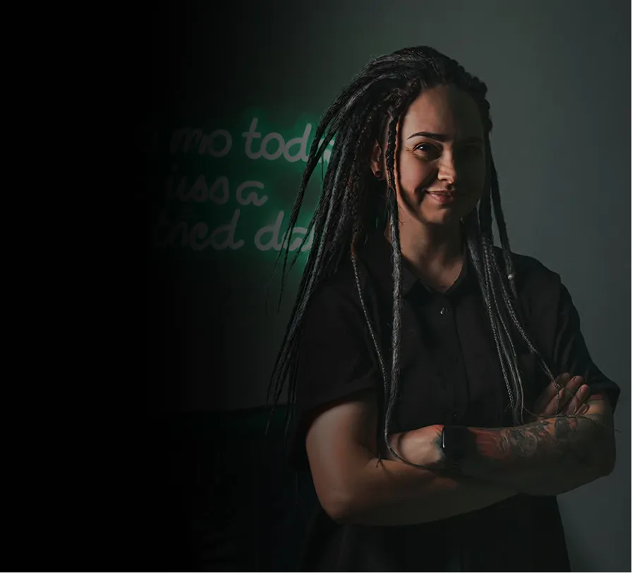Person with long dreadlocks and tattooed arm smiling with arms crossed against dark background.