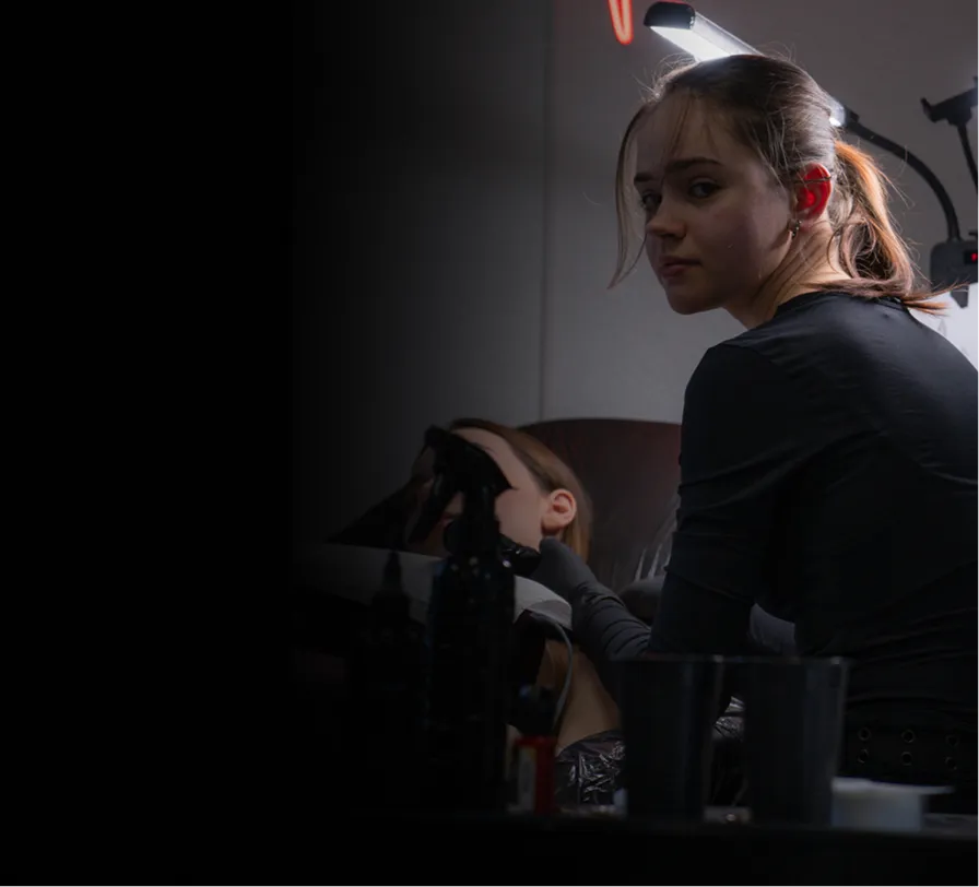 Young woman with ponytail, wearing black, looks toward camera while performing a facial treatment.