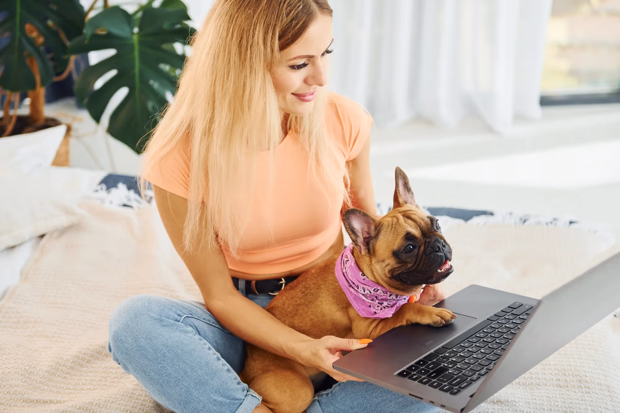 A woman sitting on a bed with a small dog on her lap and a laptop in front of her.